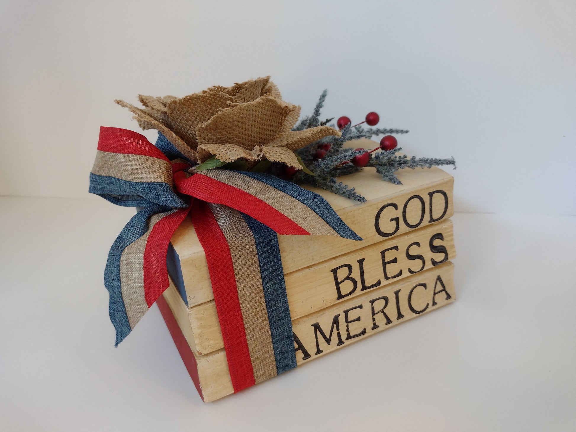 Decorative stacked books with 'God Bless America' text, red and blue ribbon, and burlap bow on a white background