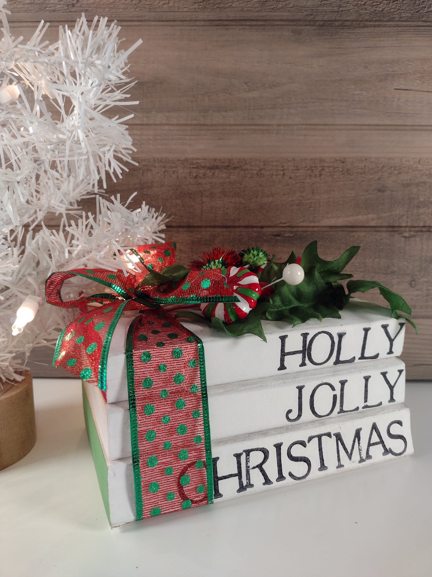 A stacked book set with a 'Holly Jolly Christmas' sign, decorated with a red and green bow and placed under a decorated Christmas tree.